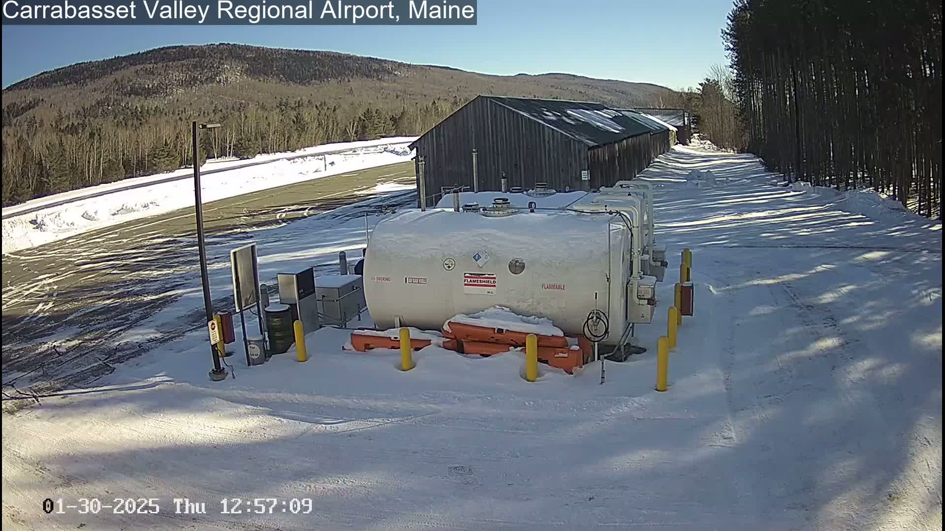 A snowy airfield with a large, white fuel tank, a dark-colored building, and a line of trees in the background under a clear, sunny sky.