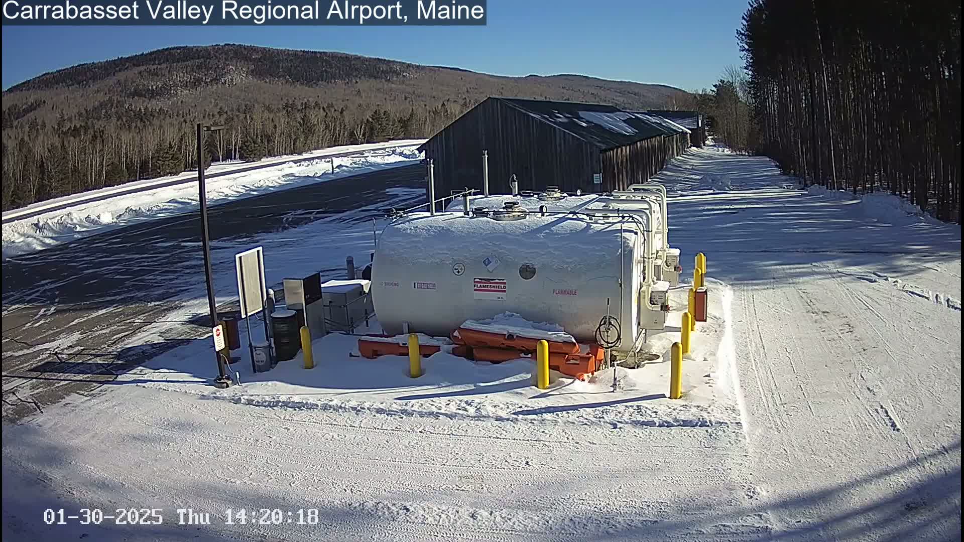 A snow-covered airport fuel depot sits in a snowy landscape under a clear, sunny sky, with a wooded mountain in the background.