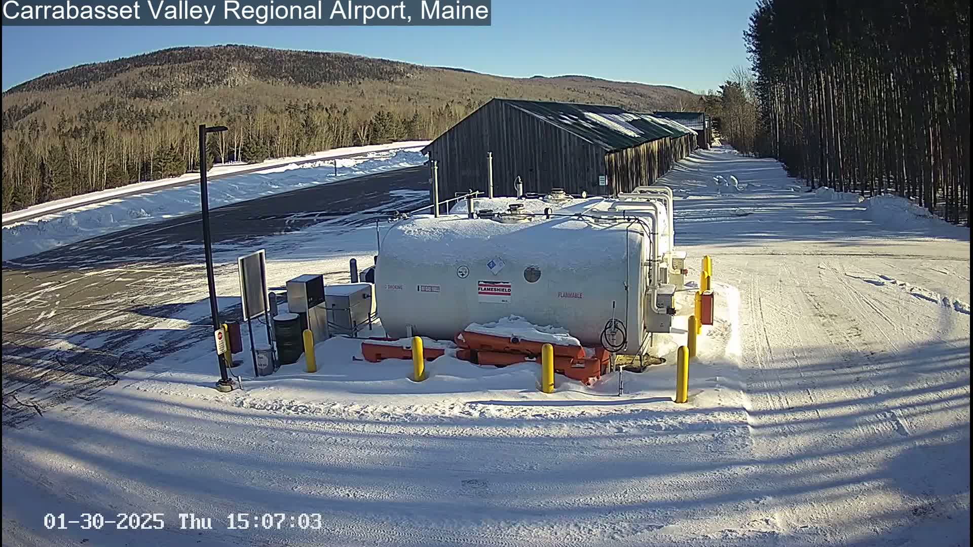 A snow-covered airport fuel depot sits near a long, dark building under a bright, sunny sky.