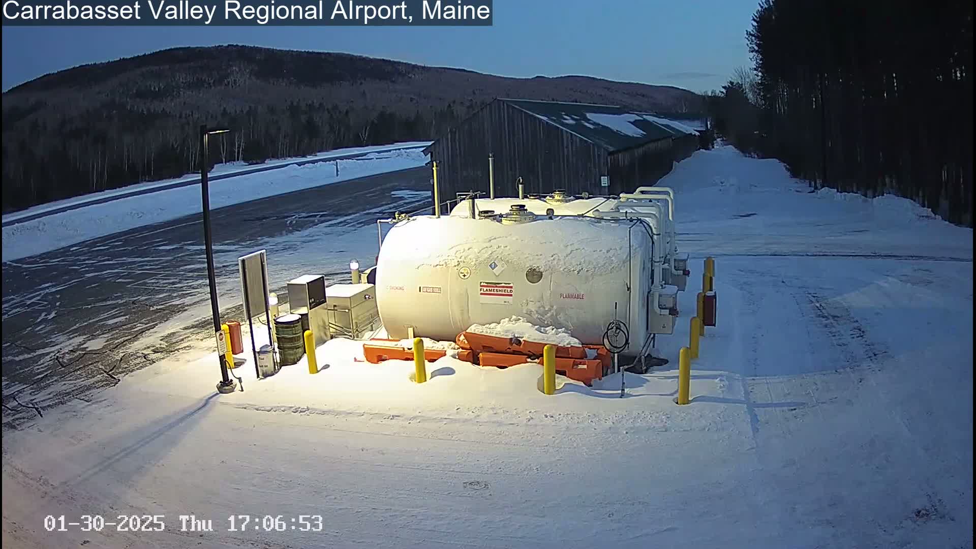 A snow-covered fuel tank sits in a snowy parking lot near a dark building, under a twilight sky.