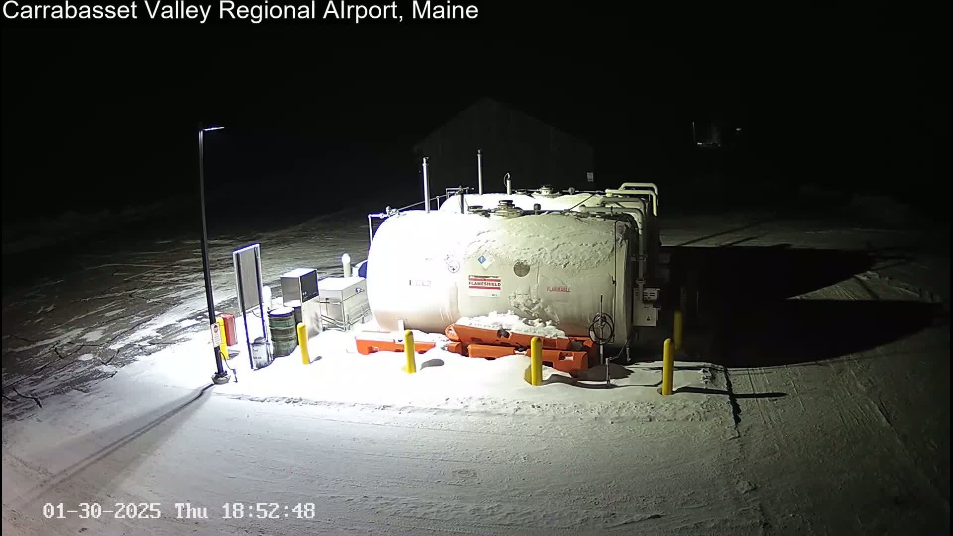 A large, snow-covered fuel tank sits in a snow-covered outdoor area at night, illuminated by a single lamp post.