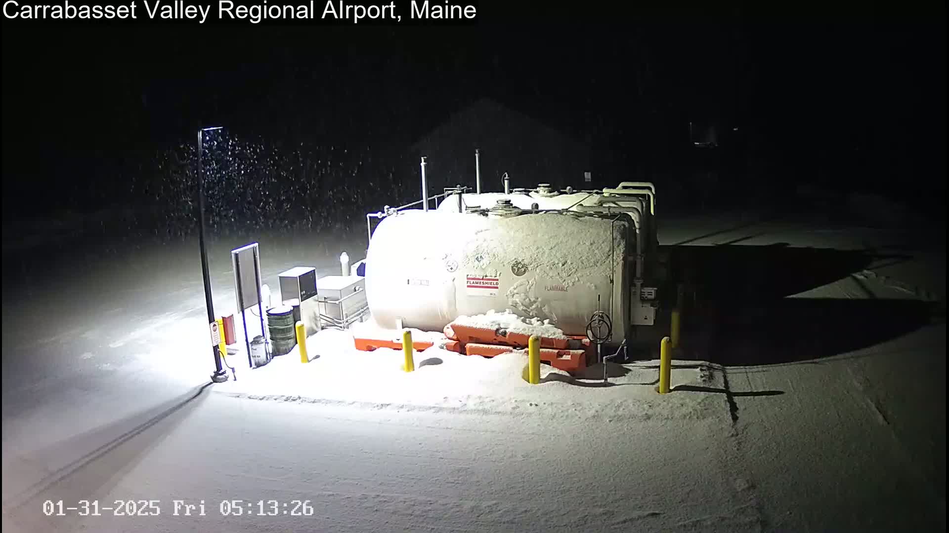 A large, snow-covered fuel tank sits in a snow-covered parking lot at night during a light snowfall.