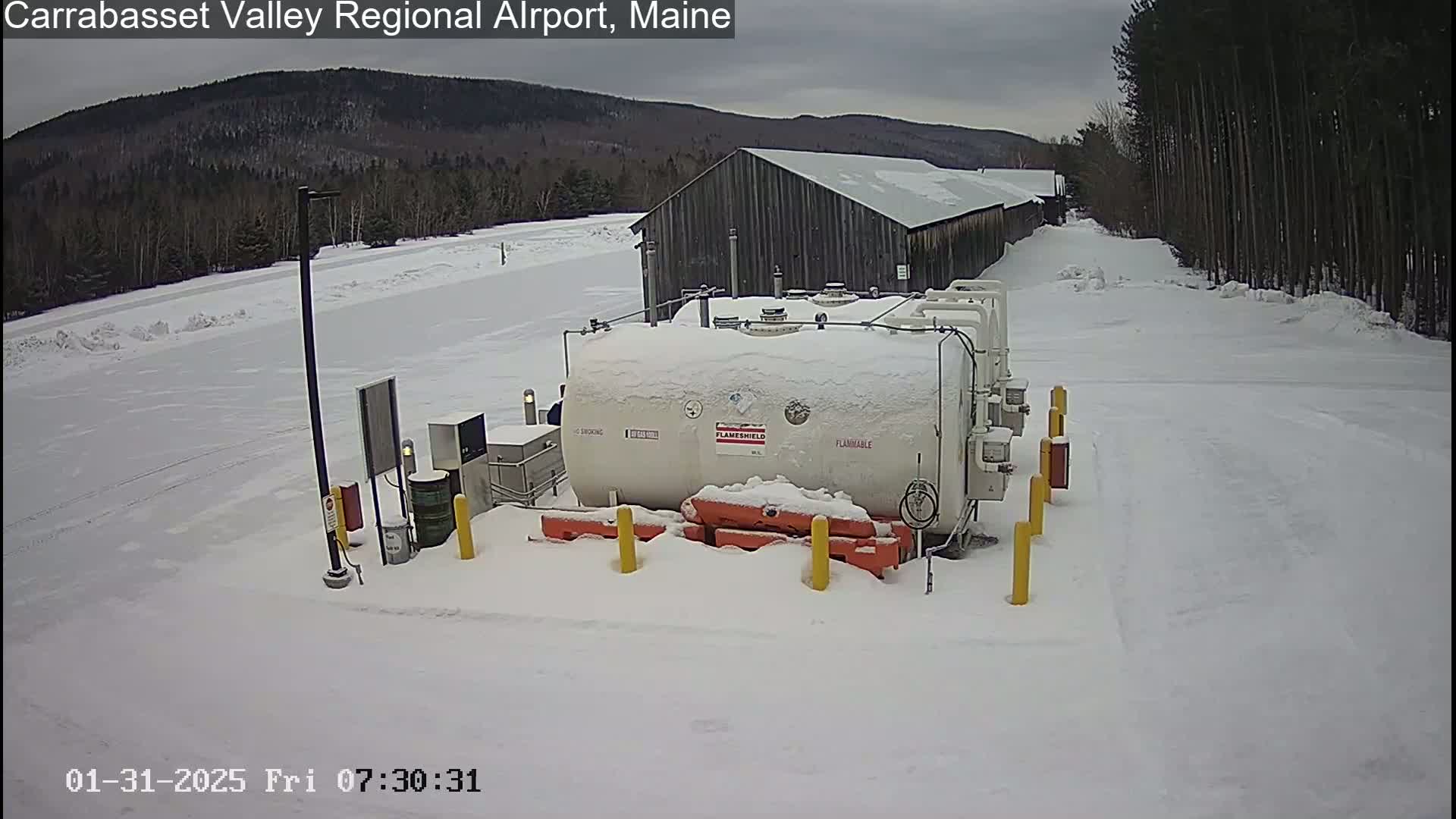 A snow-covered fuel tank sits in a snowy airport lot with a long, grey building and a snow-covered landscape in the background under an overcast sky.