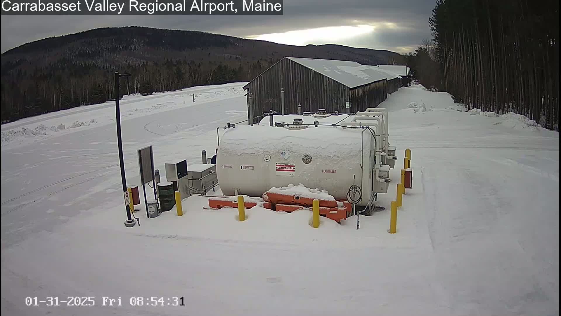 A large, snow-covered fuel tank sits in a snowy airport parking lot next to a long, wooden building, with overcast skies and a background of snow-covered trees and mountains.