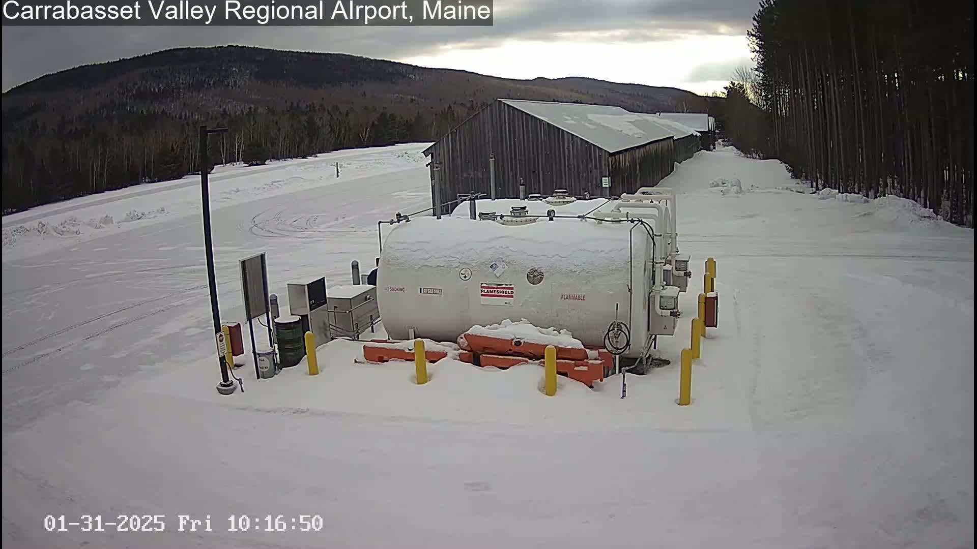 A snow-covered fuel tank sits in a snowy airfield next to a long, dark building, with a background of snow-covered trees and mountains under a cloudy sky.