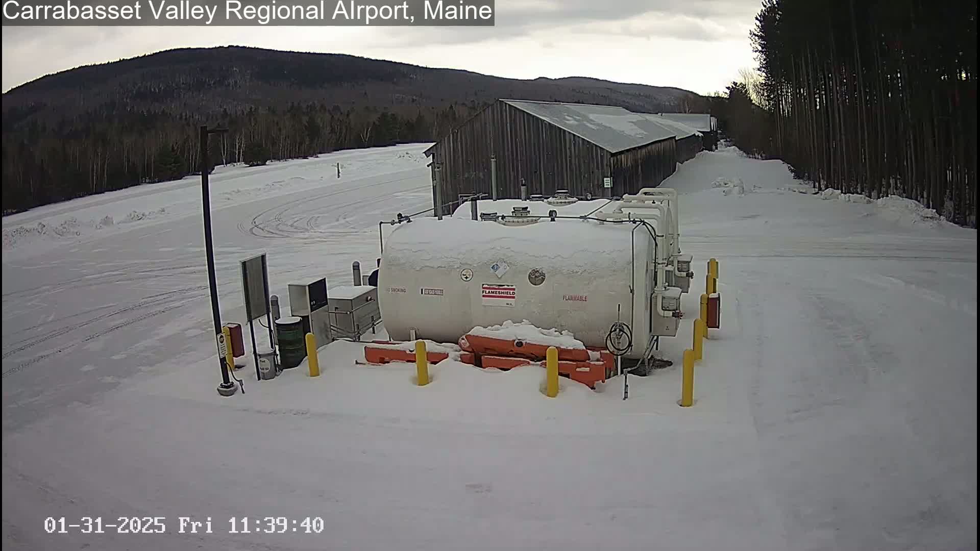 A large, snow-covered fuel tank sits in a snowy airfield next to a long wooden building, with a background of snow-covered trees and mountains under an overcast sky.