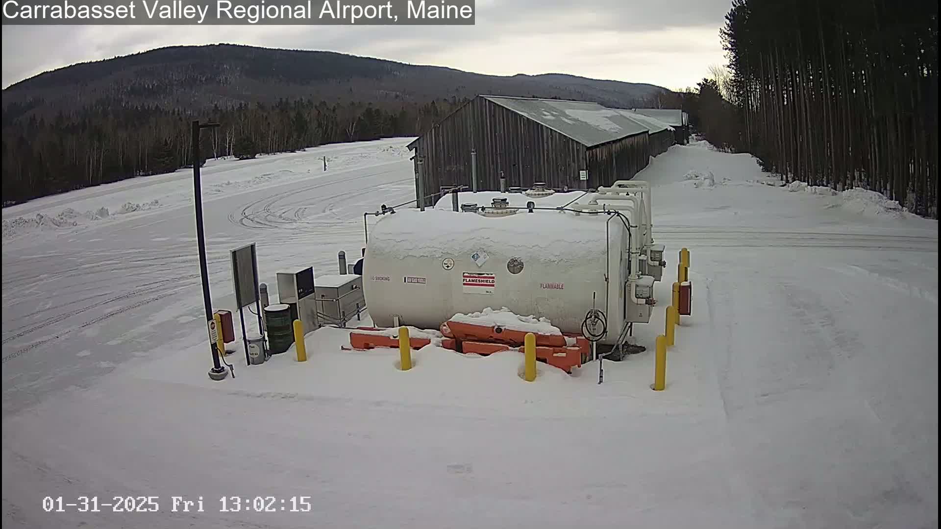 A large, snow-covered fuel tank sits in a snowy airport parking lot near a long, dark building, with a mountain range visible in the background under an overcast sky.