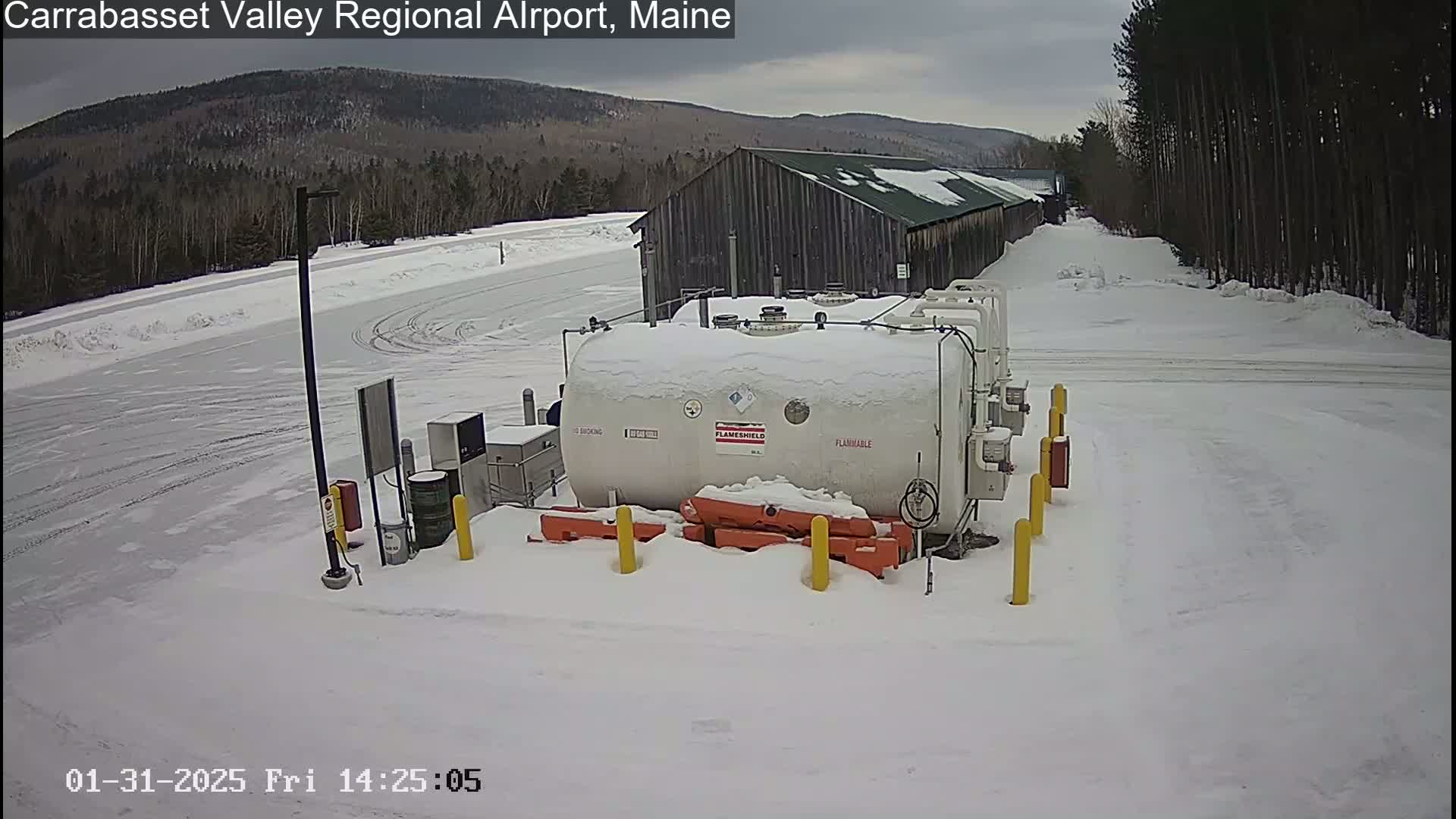 A snow-covered fuel tank sits near a long, dark-colored building in a snowy, wooded area.