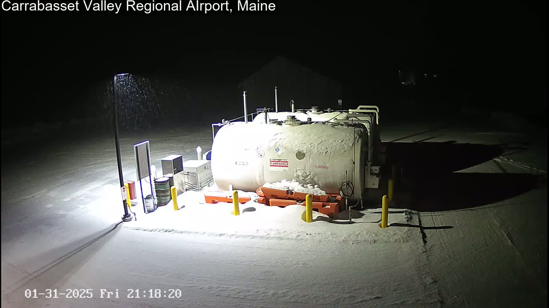 A large, snow-covered fuel tank sits in a snowy, nighttime airport lot under light snowfall.
