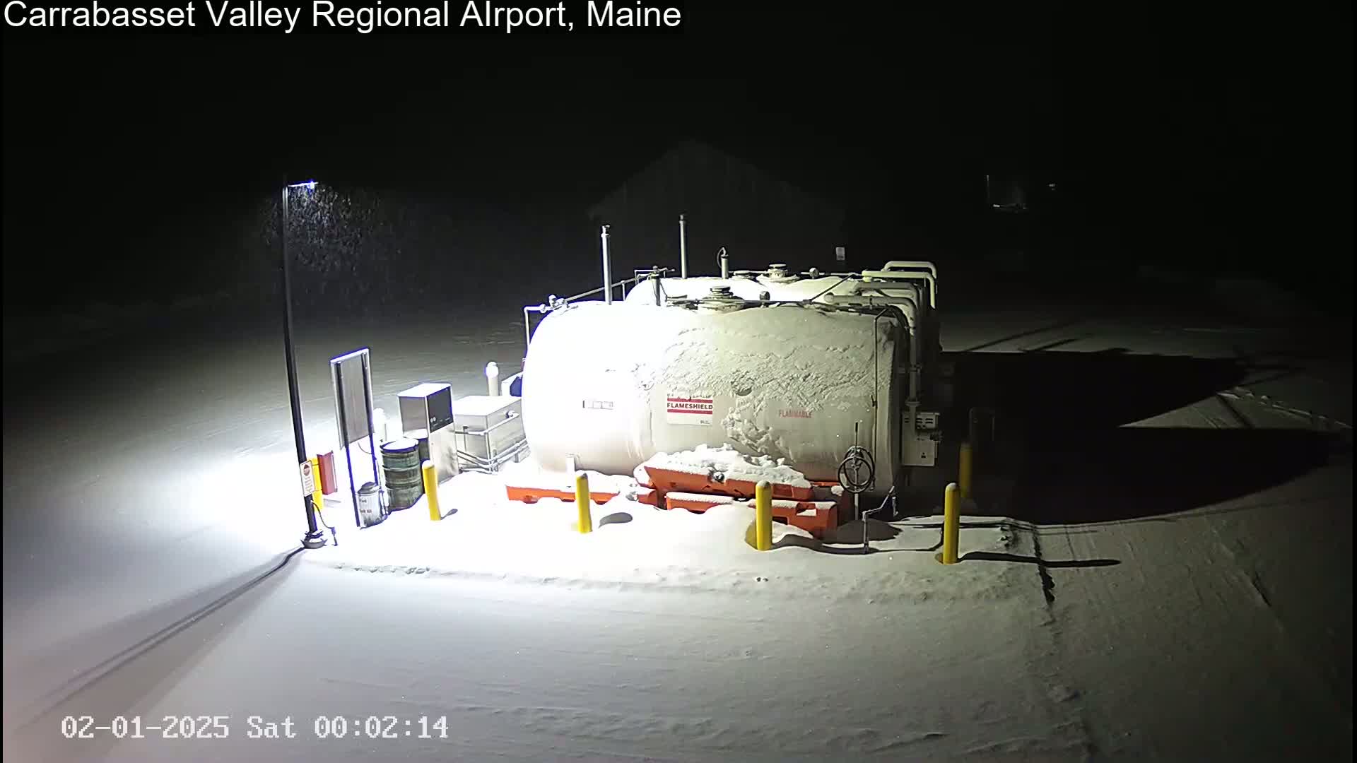 A snow-covered fuel tank and surrounding equipment are illuminated at night by a single light post during a light snowfall.