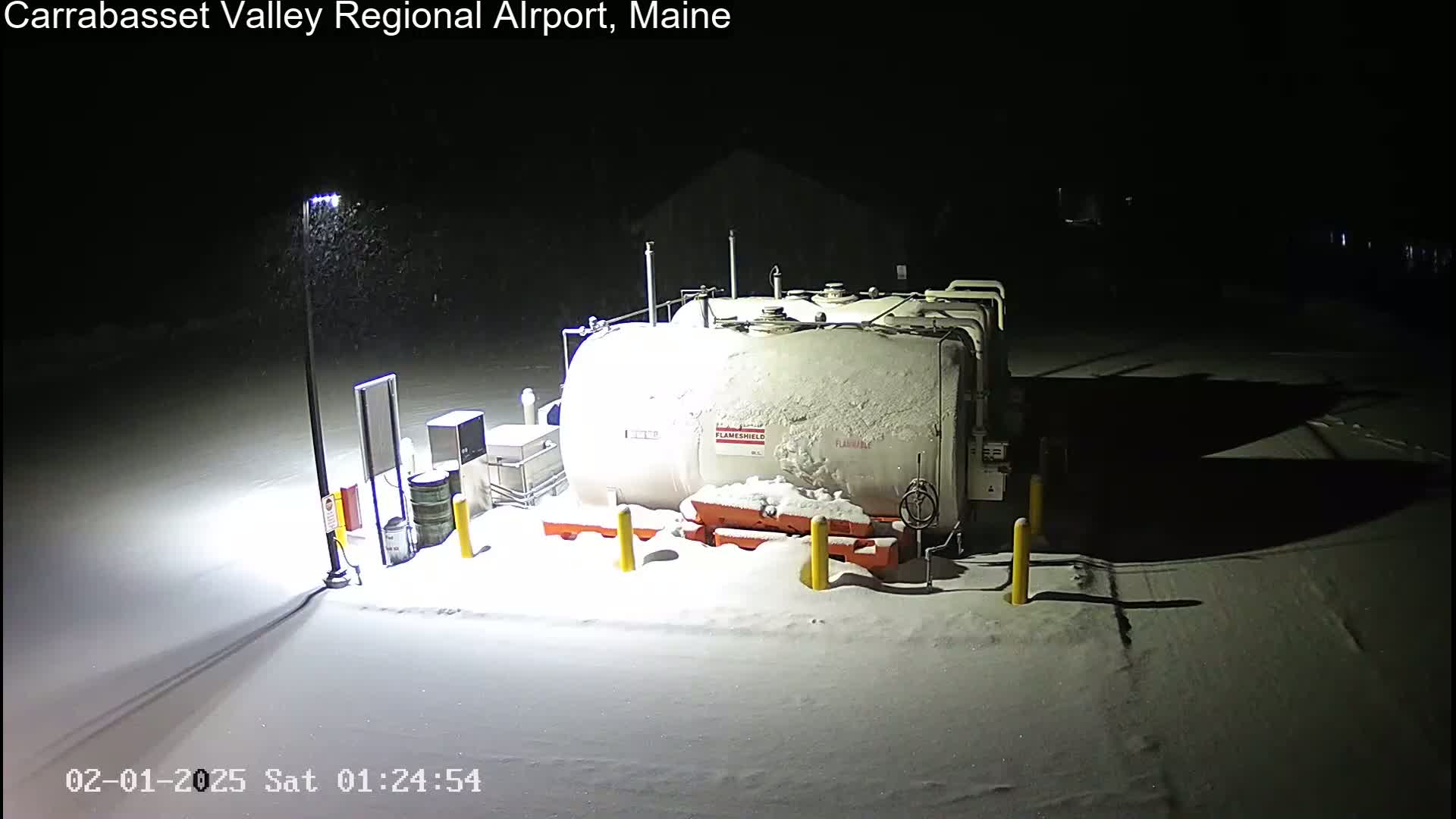A large, snow-covered fuel tank sits in a snow-covered area at night, illuminated by a single light post under light snowfall.