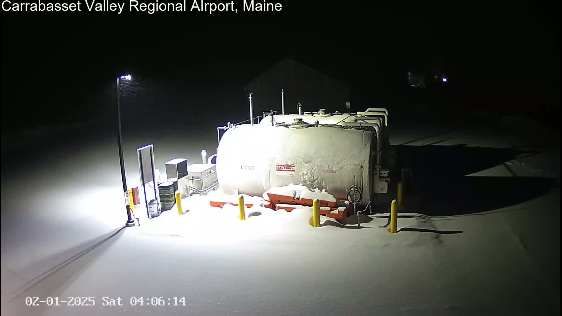 A snow-covered fuel tank and surrounding equipment are illuminated at night by a single light post during a snowfall.