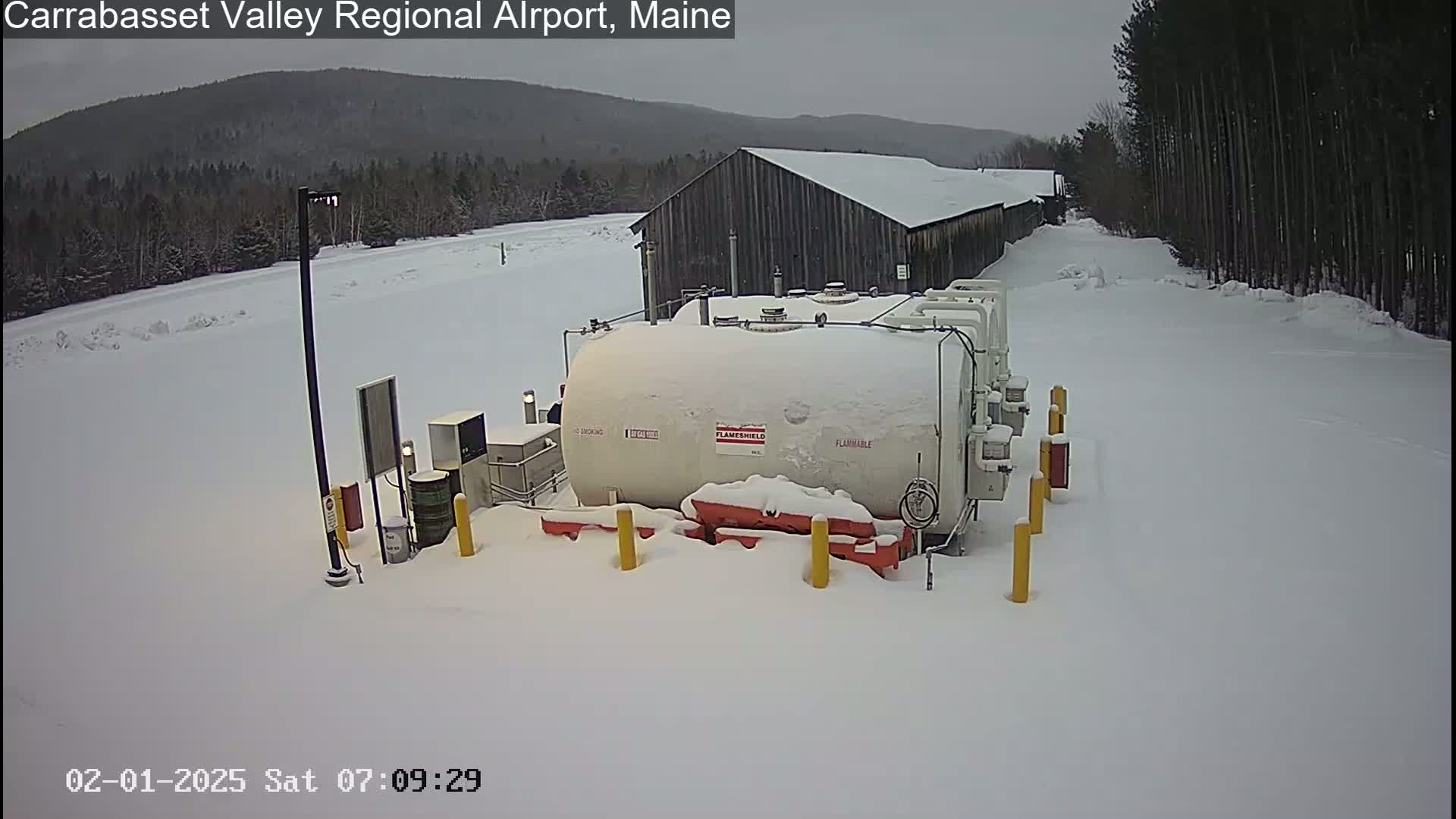 A snow-covered fuel tank sits outside a building in a snowy landscape with trees and a mountain in the background.