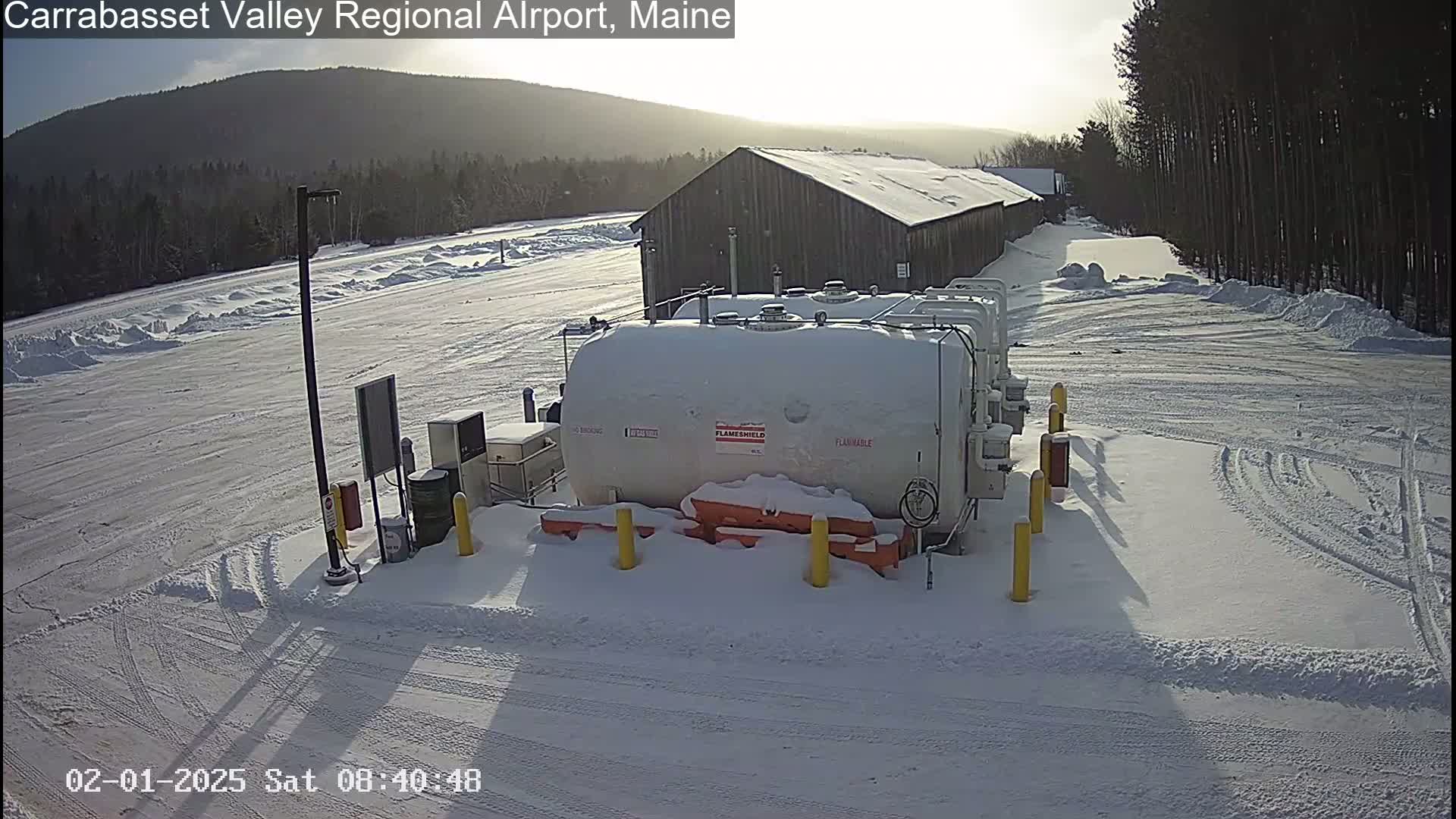 A snow-covered fuel tank farm sits in a snowy parking lot next to a long wooden building under a sunny sky.