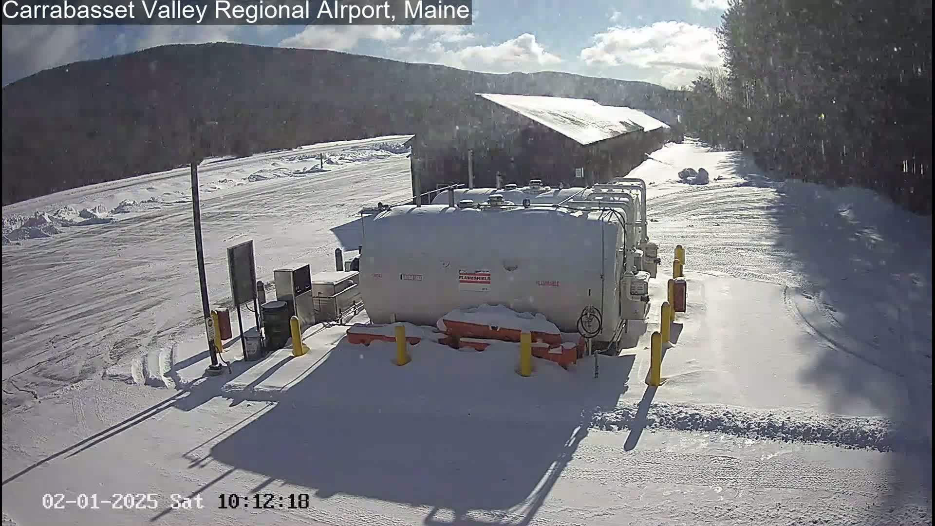 A snow-covered airport tarmac with several large white fuel tanks near a building, under a partly sunny sky with light snowfall.