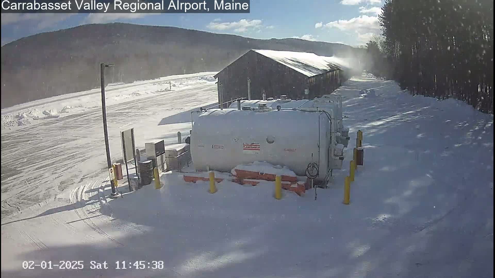 A snow-covered airport fuel farm is situated near a dark-colored building, under a partly sunny sky with light snow flurries.