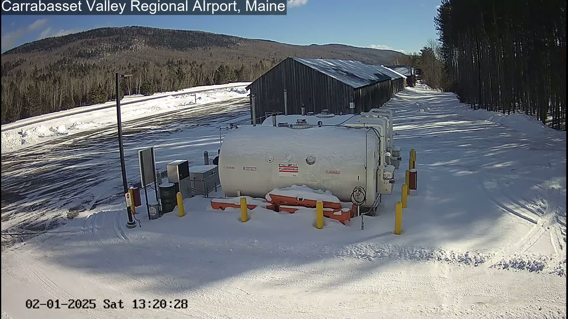 A snow-covered airport fueling station with a large tank sits next to a long, dark building, under a sunny winter sky.