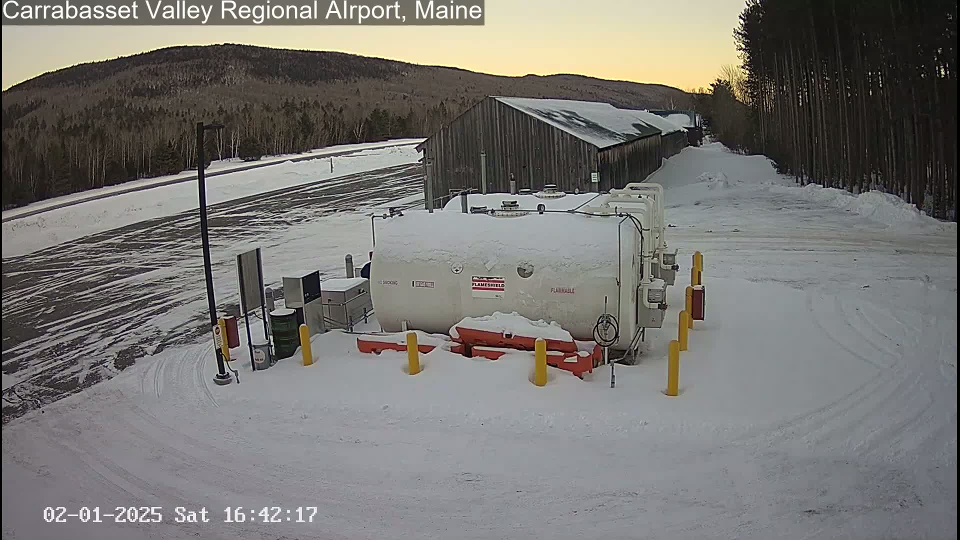 A snow-covered airport fuel storage area with a large white tank and a long, gray building in the background under a twilight sky.