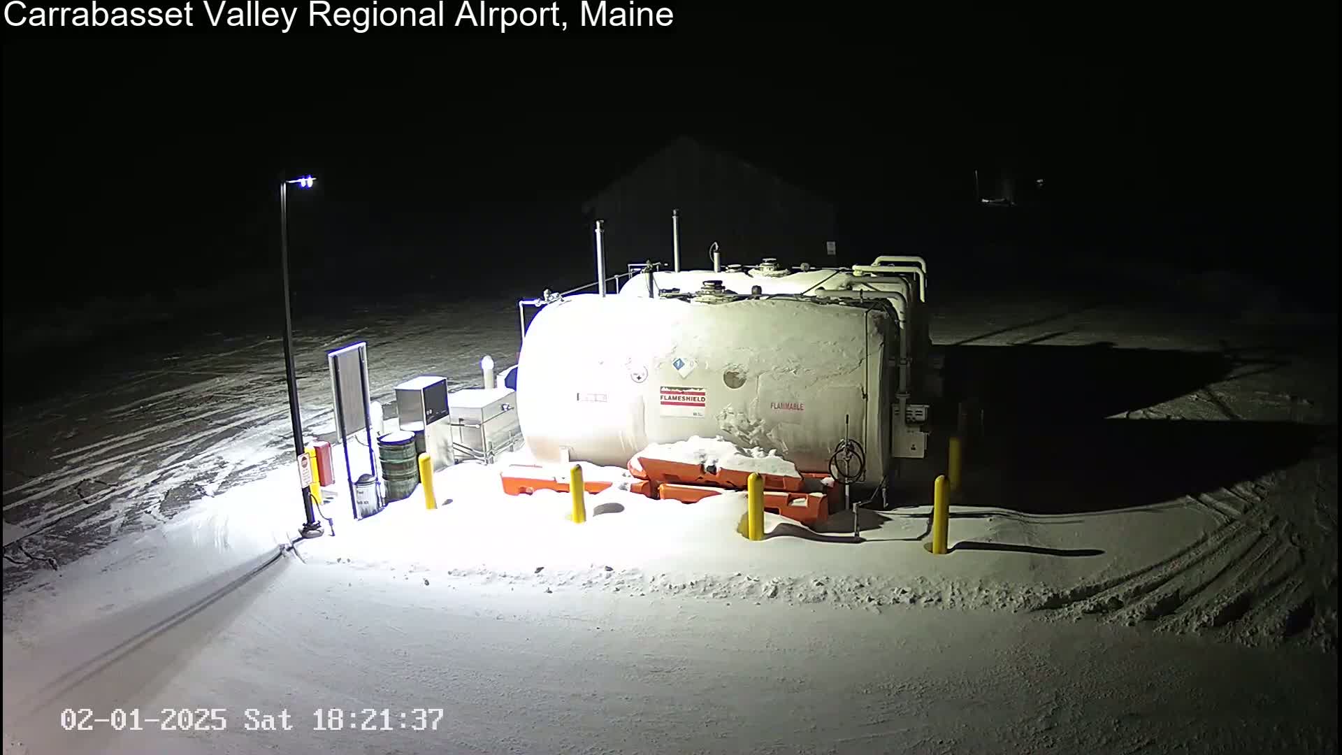 A large, snow-covered fuel tank sits in a snow-covered lot at night, illuminated by a single light pole.
