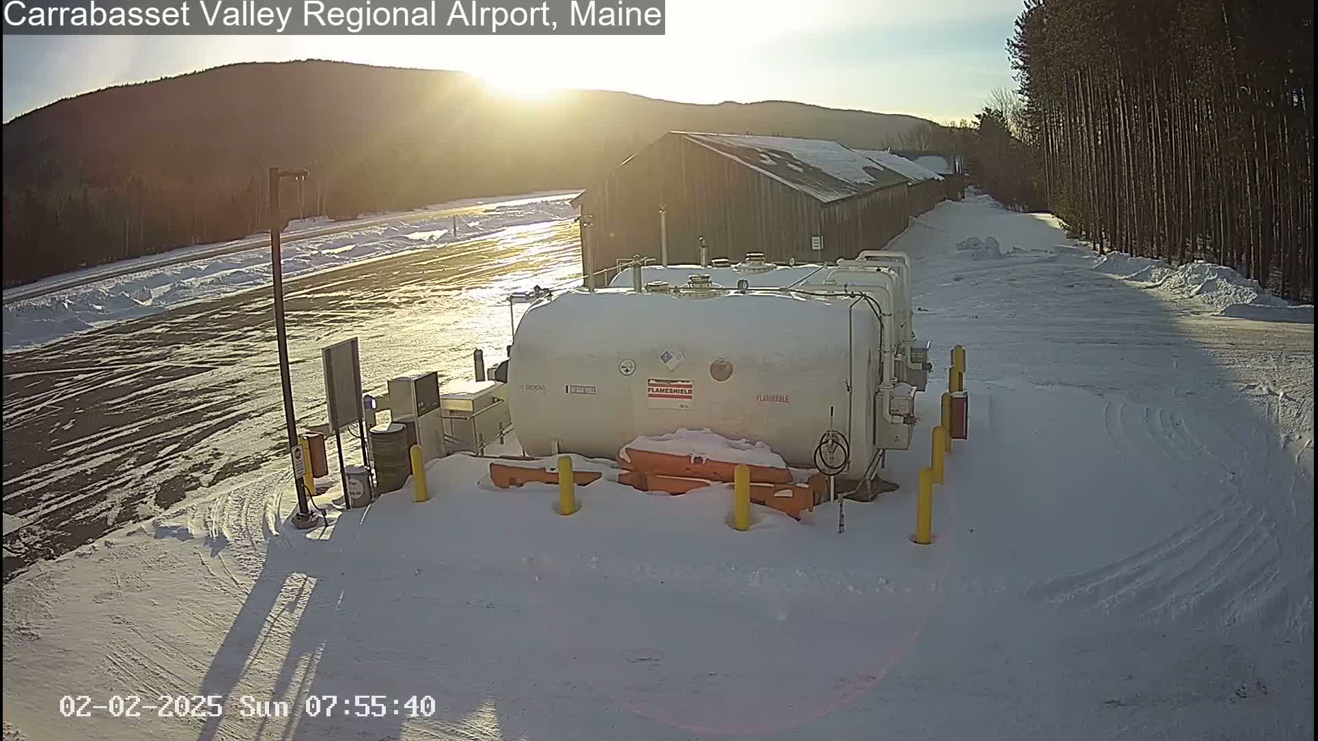 A large, white, flammable liquid storage tank sits in a snowy airfield parking lot, with a barn-like structure visible in the background under a bright, sunny sky.