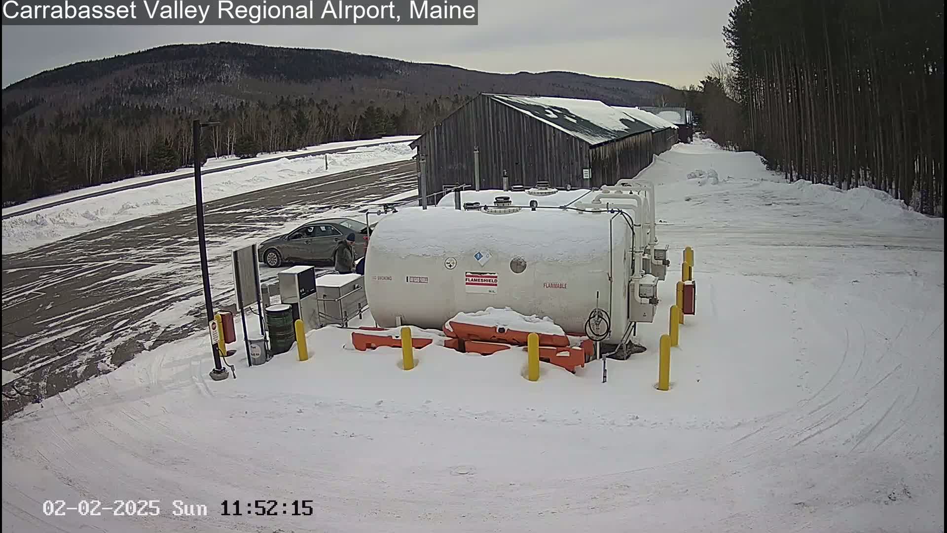 A snow-covered airport parking lot contains a large white fuel tank, a car, and a long, low building in the background under an overcast sky.