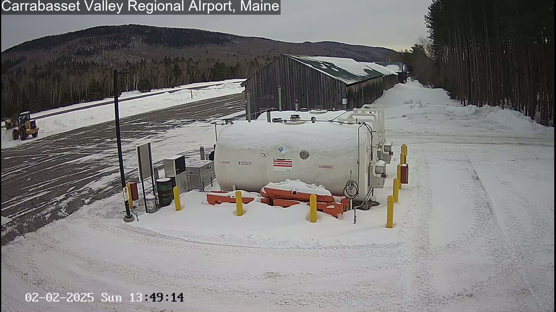 A snow-covered airport tarmac features a large white fuel tank near a long, dark building, with a yellow piece of heavy machinery visible in the distance under a cloudy sky.
