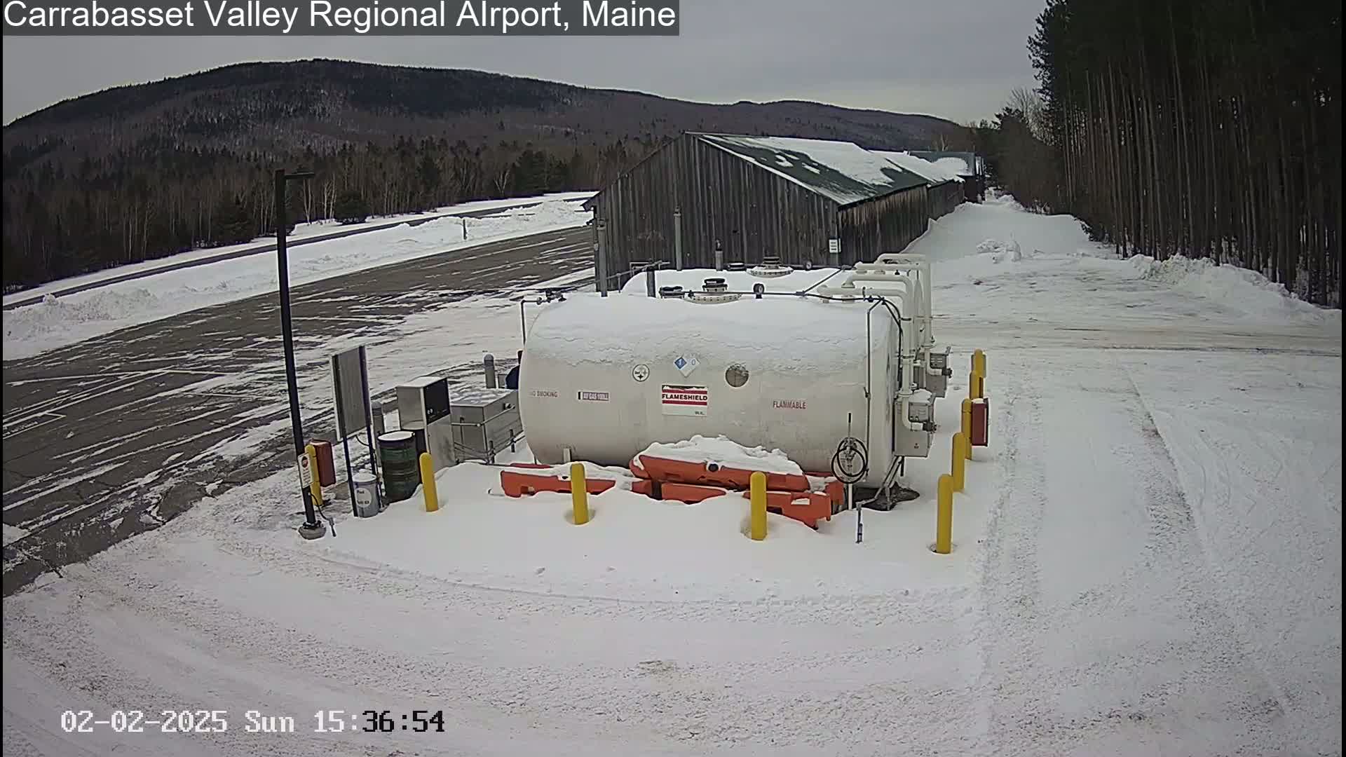 A large, white, cylindrical tank labeled "flammable" sits in a snow-covered parking lot near a long, gray building, with a wooded hillside visible in the background under an overcast sky.