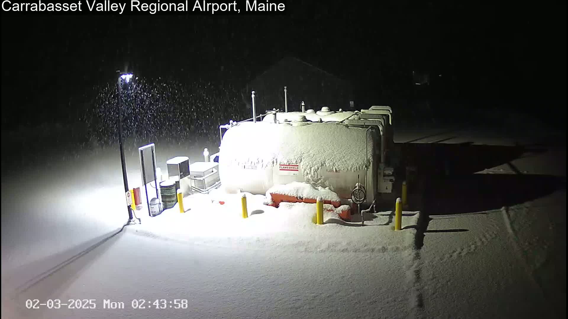 A snow-covered fuel tank and surrounding equipment are illuminated by a lamp post during a nighttime snowfall.