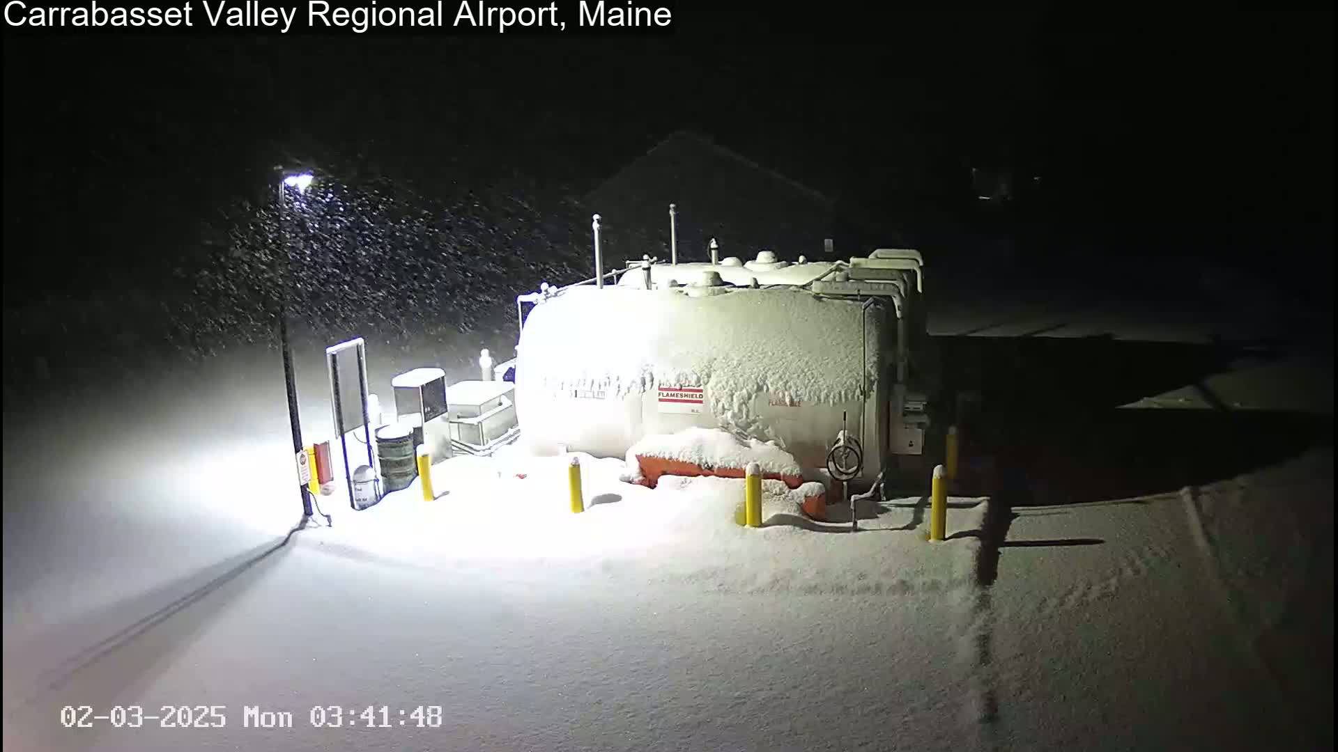 A large, snow-covered fuel tank and surrounding equipment are illuminated by a streetlight during a nighttime snowfall.