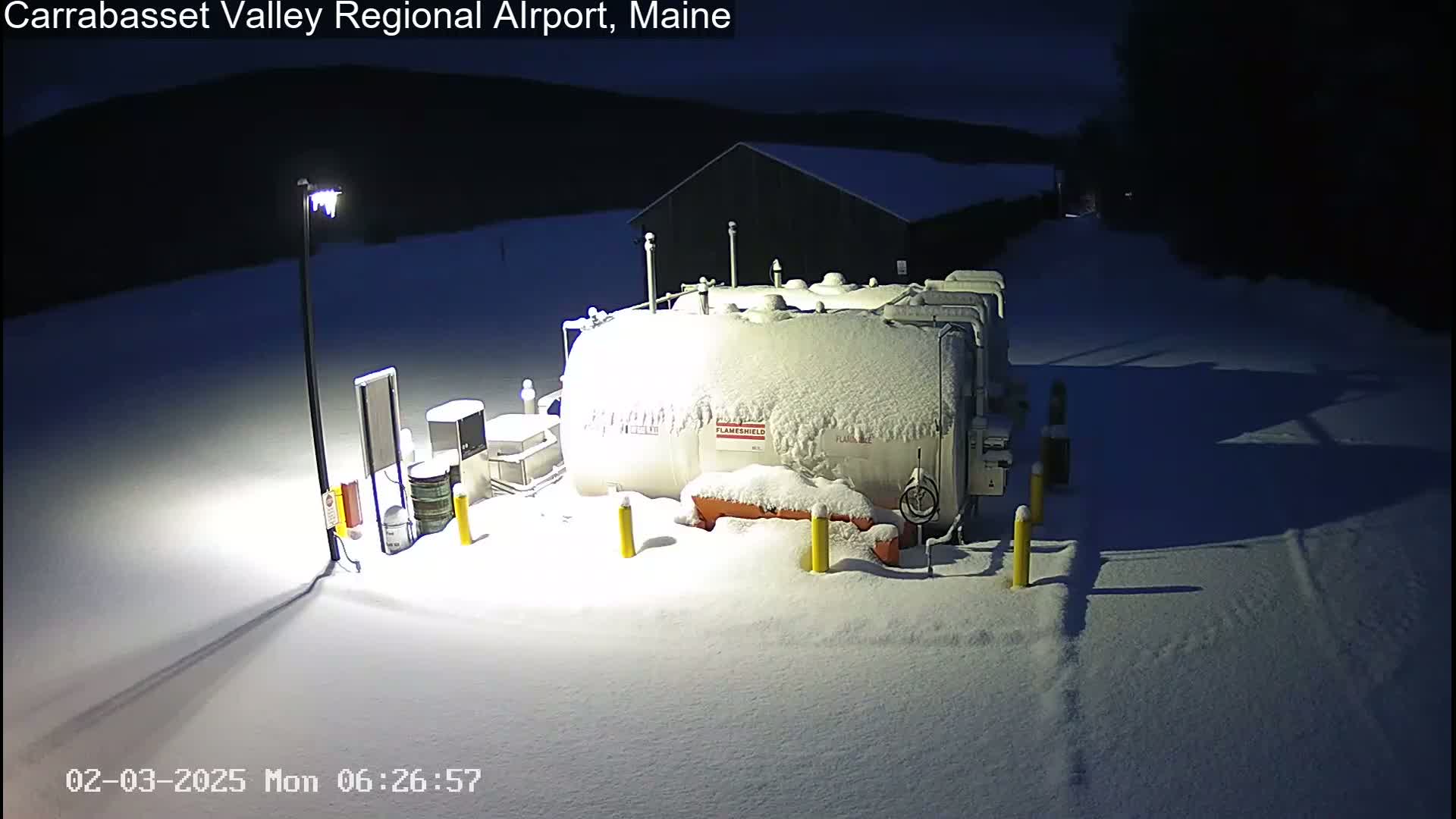 A snow-covered fuel tank and surrounding equipment are illuminated at night under a dark sky.
