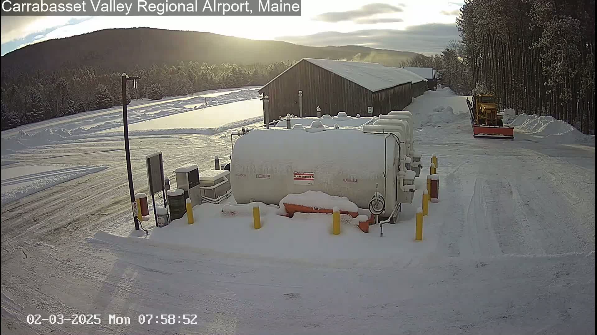 A snow-covered airport tarmac with a large fuel tank, a snowplow, and a long wooden building in the background under a partly sunny sky.