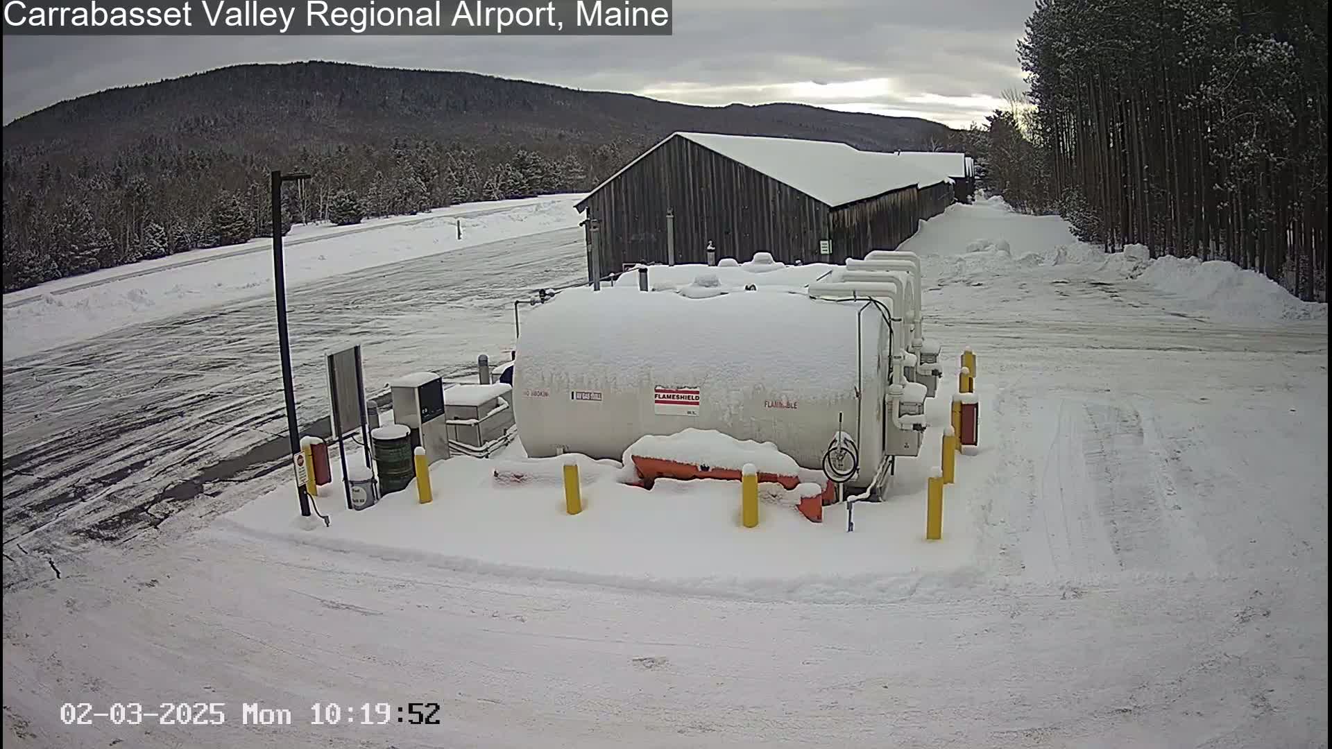 A snow-covered fuel tank sits in a snowy airport parking lot near a snow-covered building and a stand of trees under a cloudy sky.