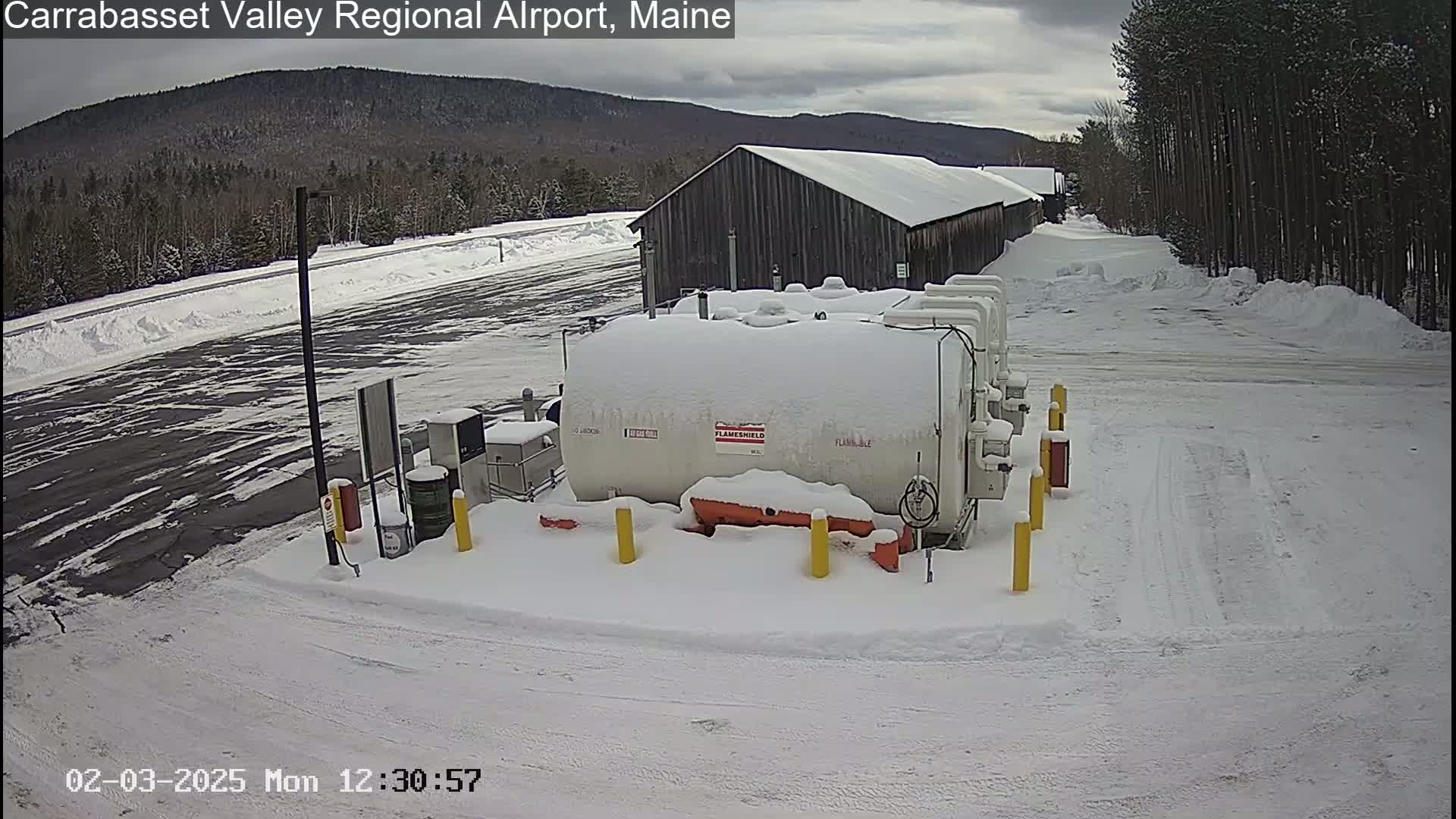 A snow-covered airport fueling station with several tanks sits near a long, snow-covered building, all under a cloudy sky.