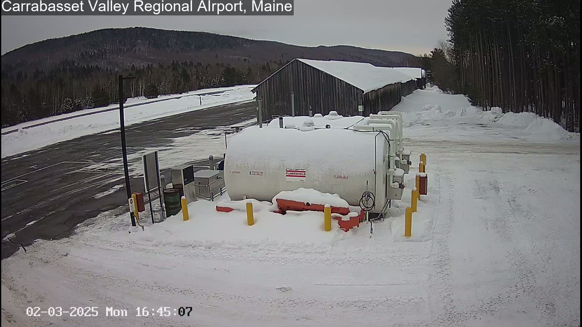 A snow-covered fuel tank sits in a snowy parking lot next to a snow-covered building, with a treeline and distant mountains in the background under a cloudy sky.
