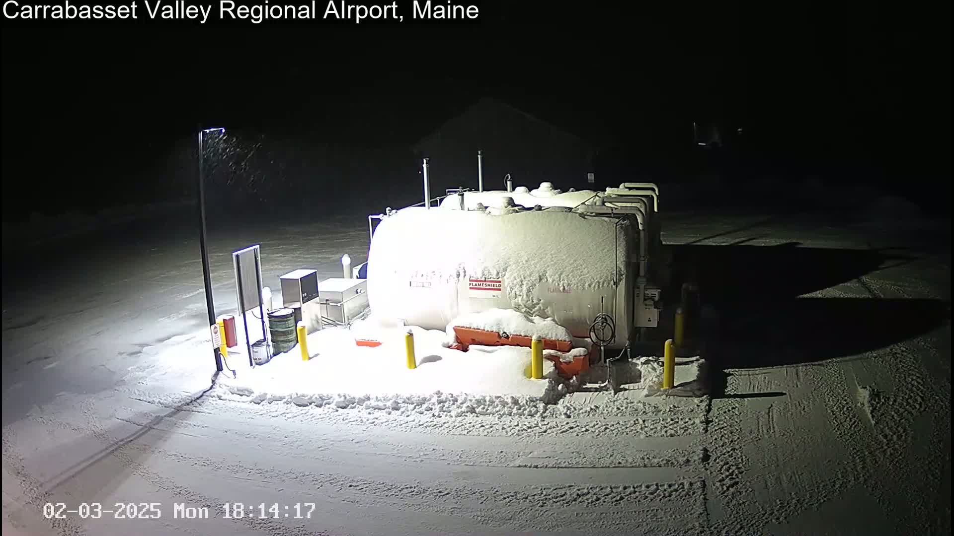 A large, snow-covered fuel tank sits in a snow-covered parking lot at night, under light snowfall.