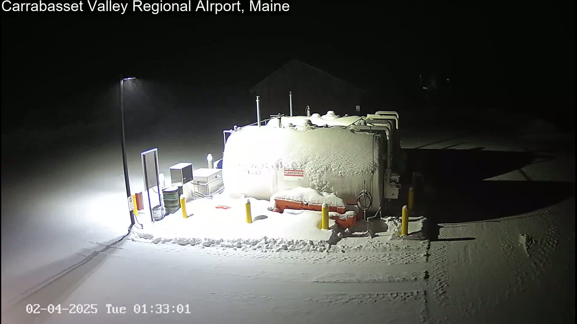 A large, snow-covered fuel tank and surrounding equipment are illuminated at night in a snowy area.