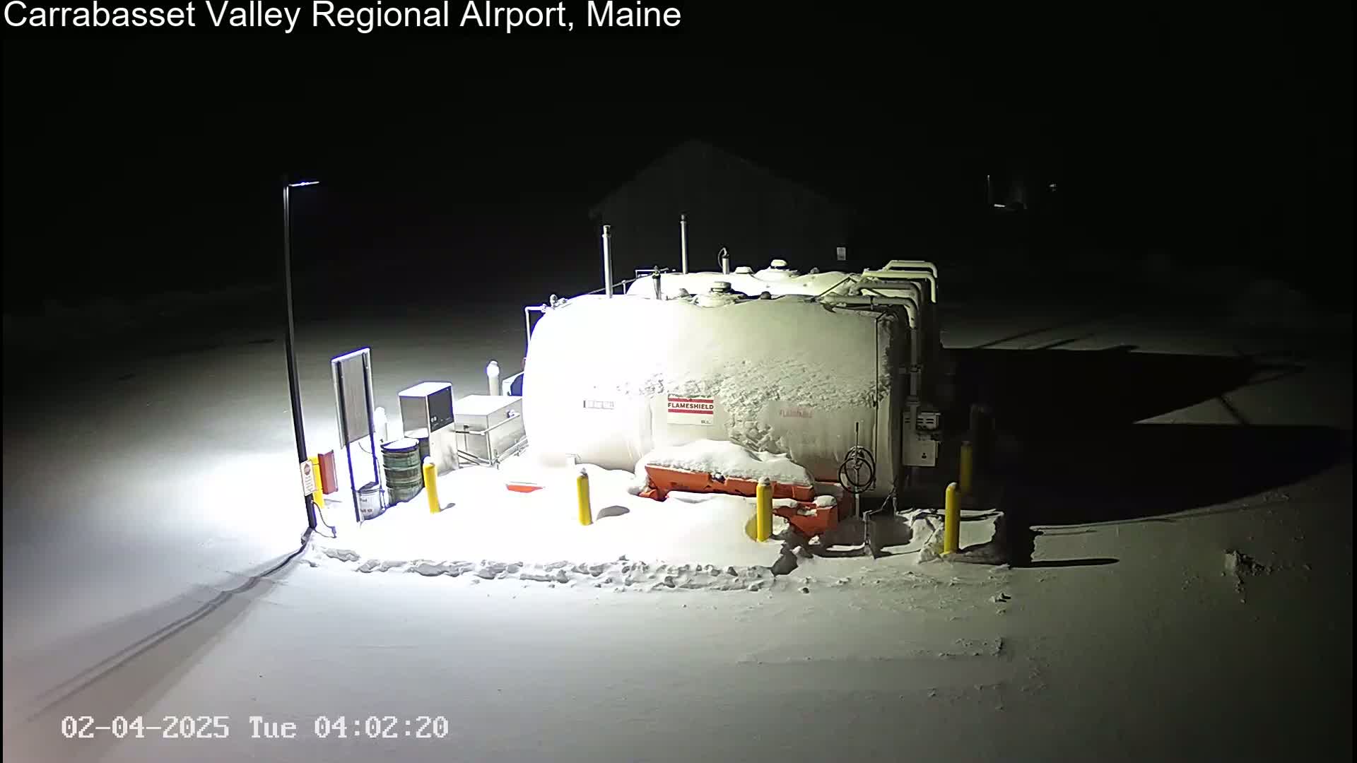 A large, snow-covered tank and surrounding equipment are illuminated at night in a snowy area.