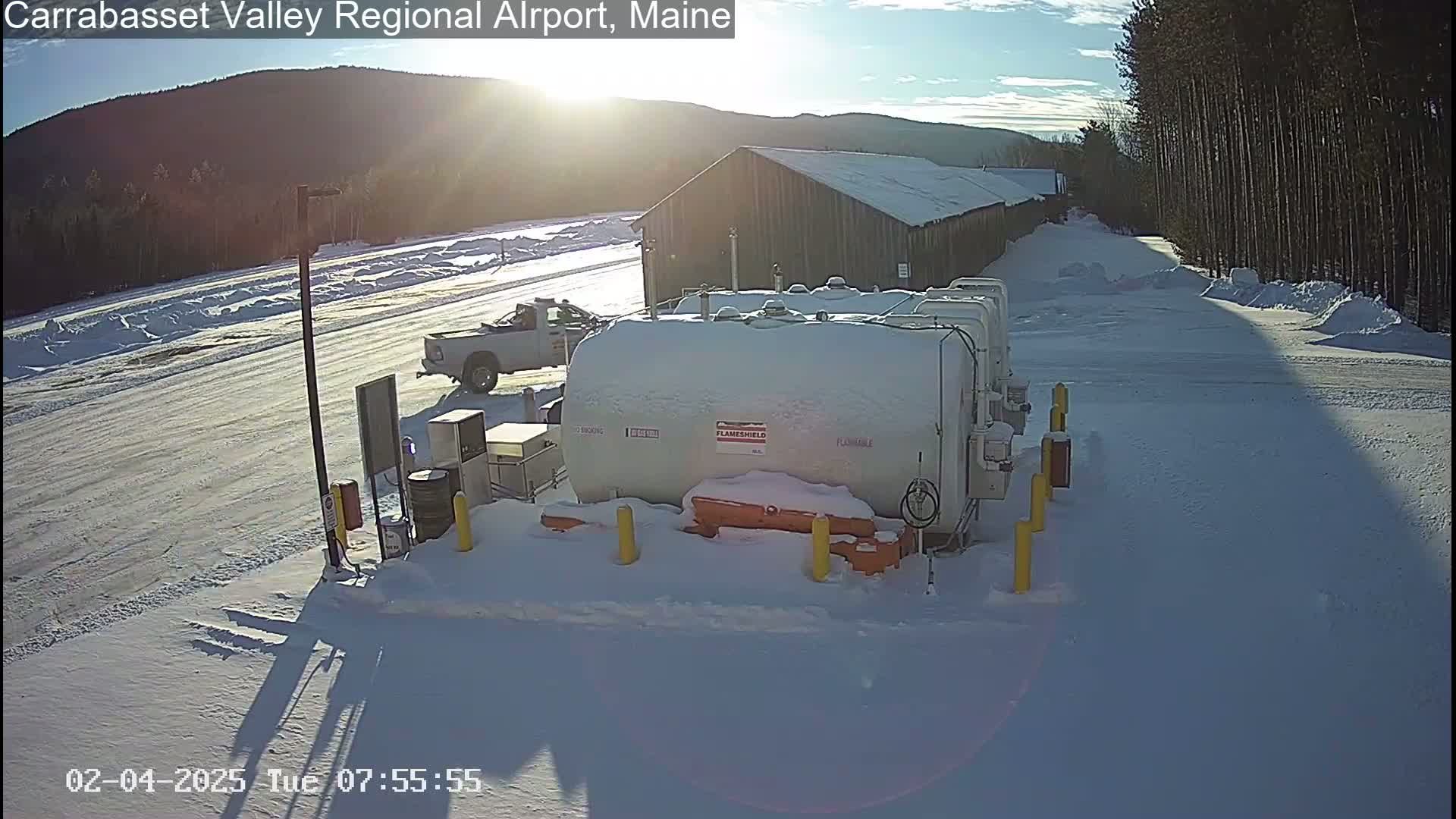 A snow-covered fuel storage area with a pickup truck parked nearby, situated on a snowy airfield under a sunny sky.