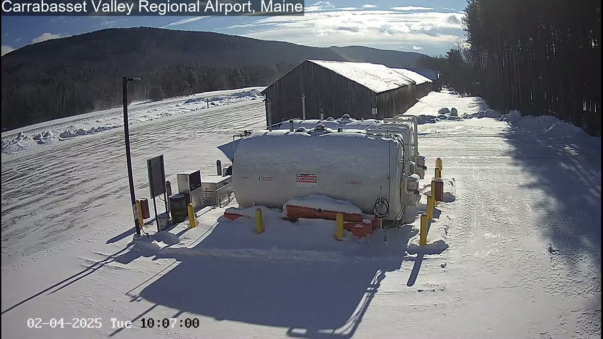 A snowy airfield scene shows several large, snow-covered fuel tanks near a long, dark building under a partly sunny sky.