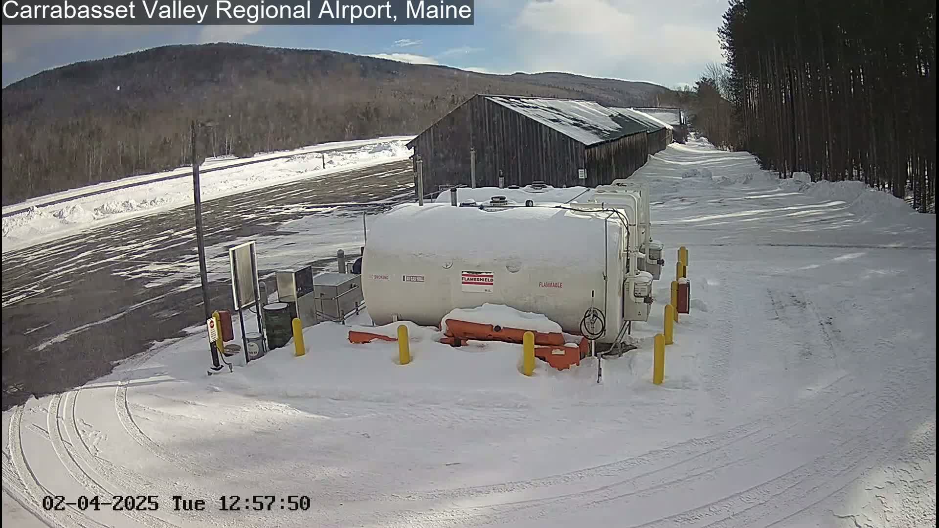 A snow-covered fuel storage area with a large white tank, a wooden barn, and tire tracks in the snow under a partly sunny sky.