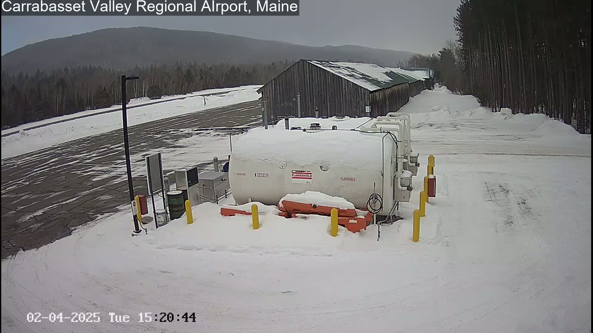A large white fuel tank covered in snow sits in a snow-covered airport parking lot near a wood building, with a wooded area and distant mountains in the background under an overcast sky.