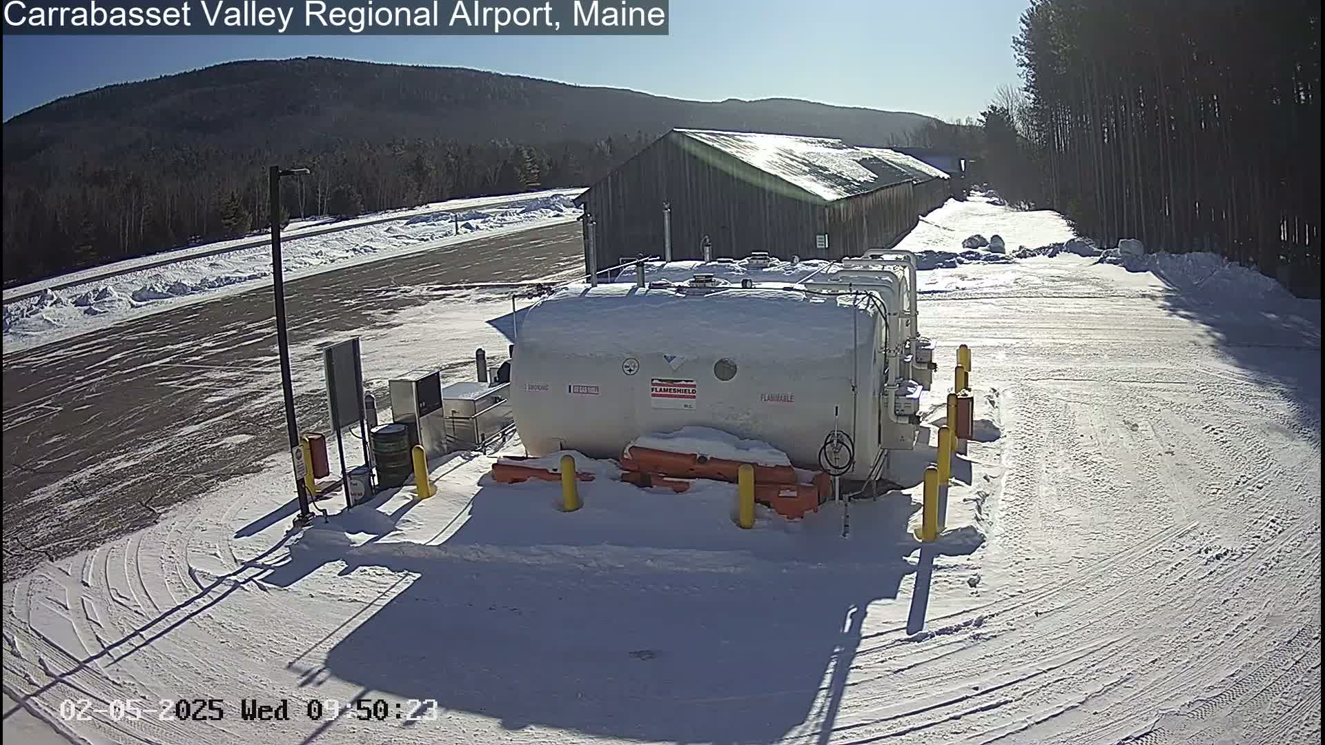 A large, snow-covered fuel tank sits in a snowy airport parking lot under a sunny sky, with a long building and distant mountains visible in the background.
