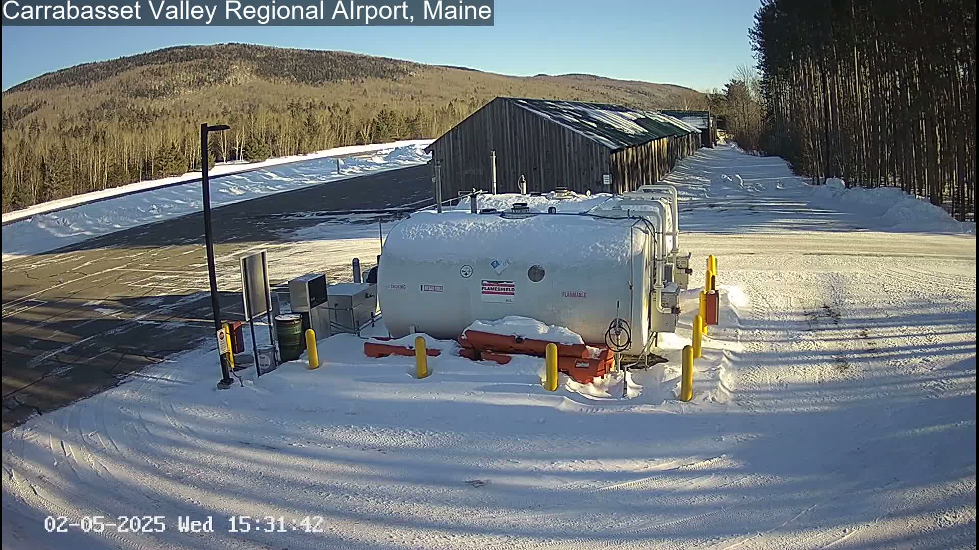 A snow-covered airport fueling station sits in a large, snow-covered parking lot on a sunny day, with a wooded hillside visible in the background.