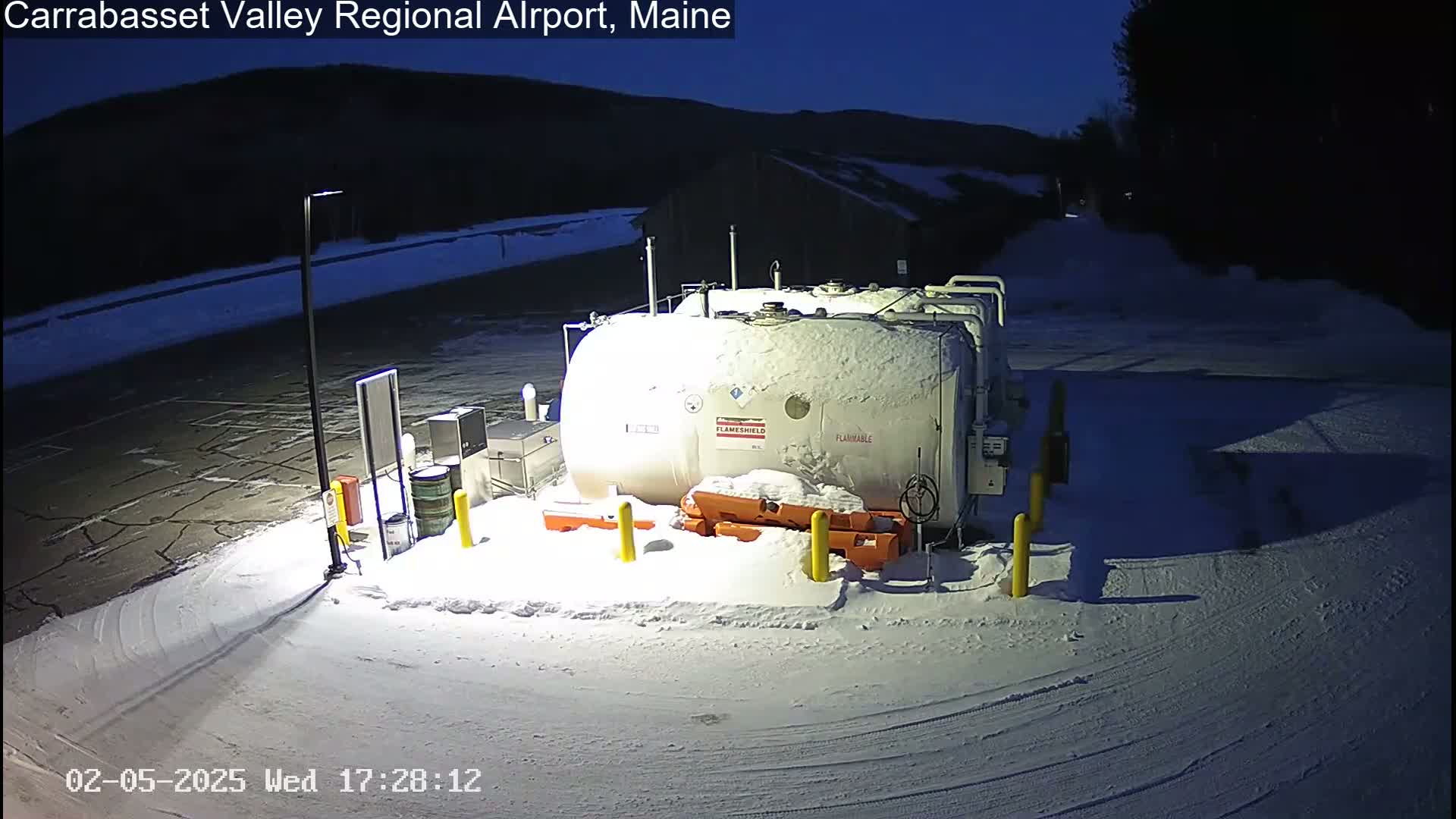 A large, snow-covered fuel tank sits in a snow-covered parking lot at night, with dark mountains in the background.