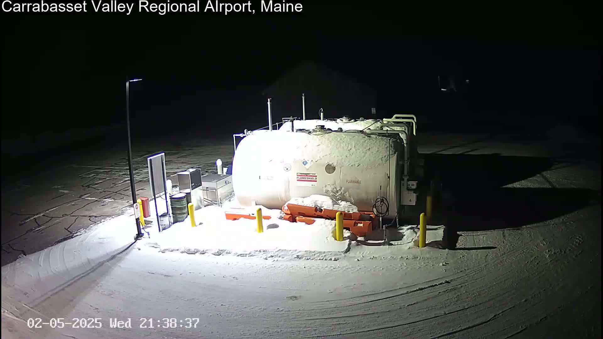 A large, snow-covered fuel tank sits in a snow-covered airport parking lot at night.