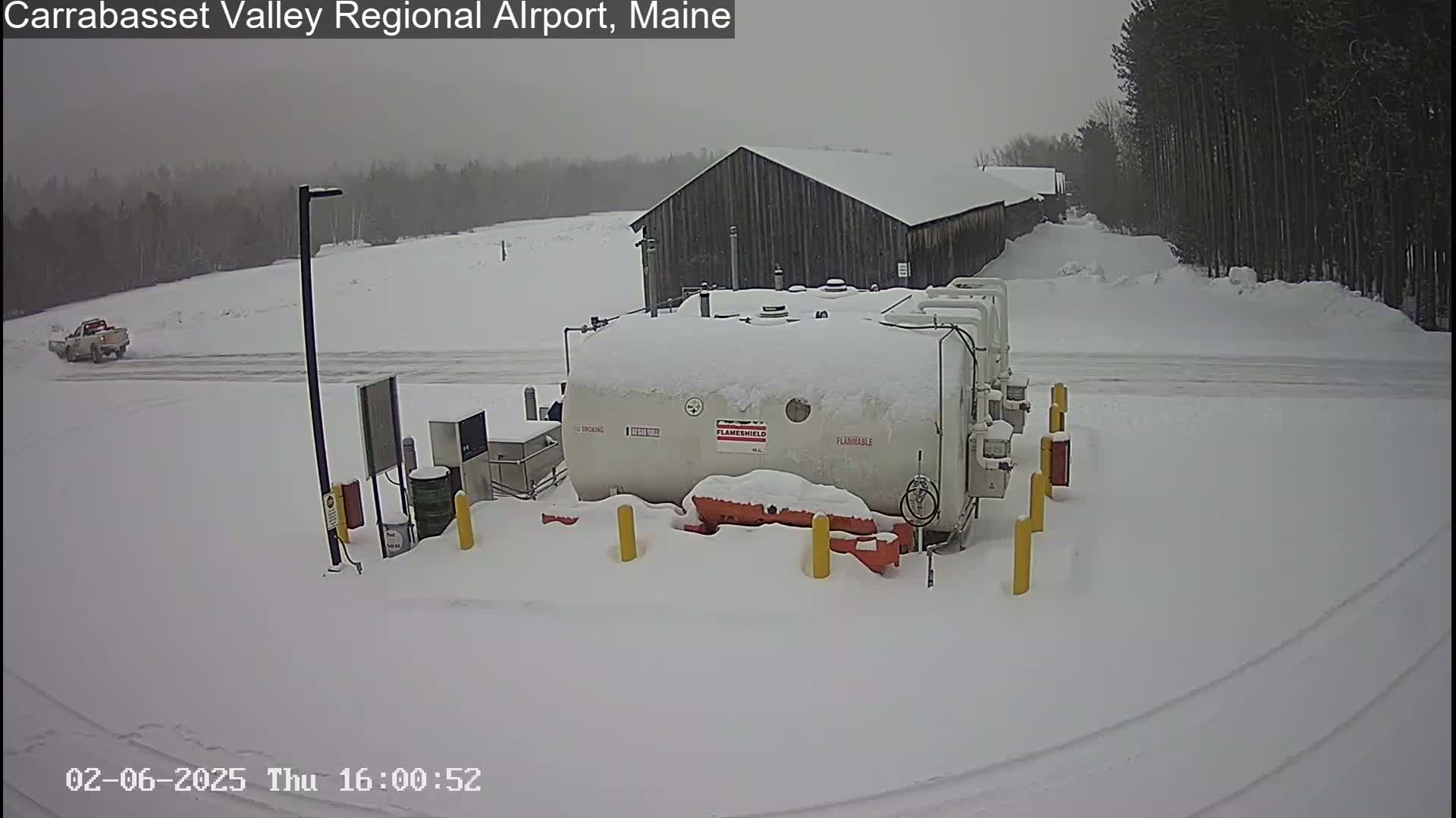 A snow-covered fuel tank sits in a snowy airfield parking lot near a snow-covered building and a truck.
