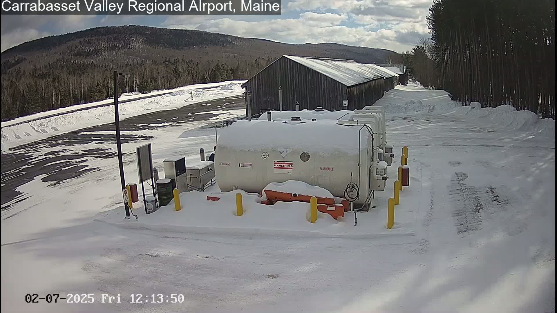 A snow-covered airport fuel farm is situated near a long, dark building, with a partly sunny sky and snow-covered ground in the background.