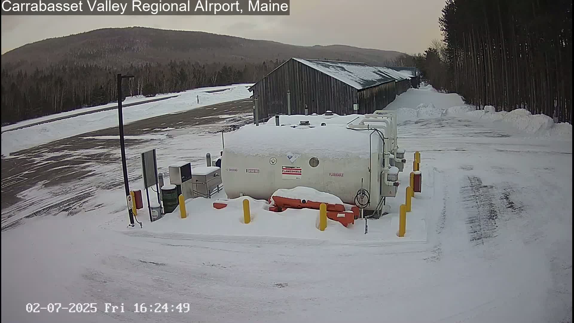 A snow-covered airport fueling station sits near a long, wooden building, with a background of snow-covered trees and distant mountains under an overcast sky.