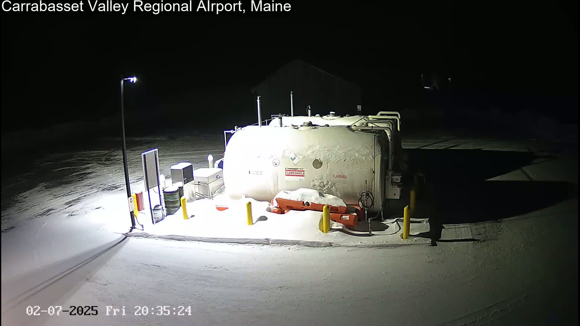 At night, under a snowy, dark sky, two large, snow-covered fuel tanks sit in a snow-covered area, illuminated by a single lamppost.