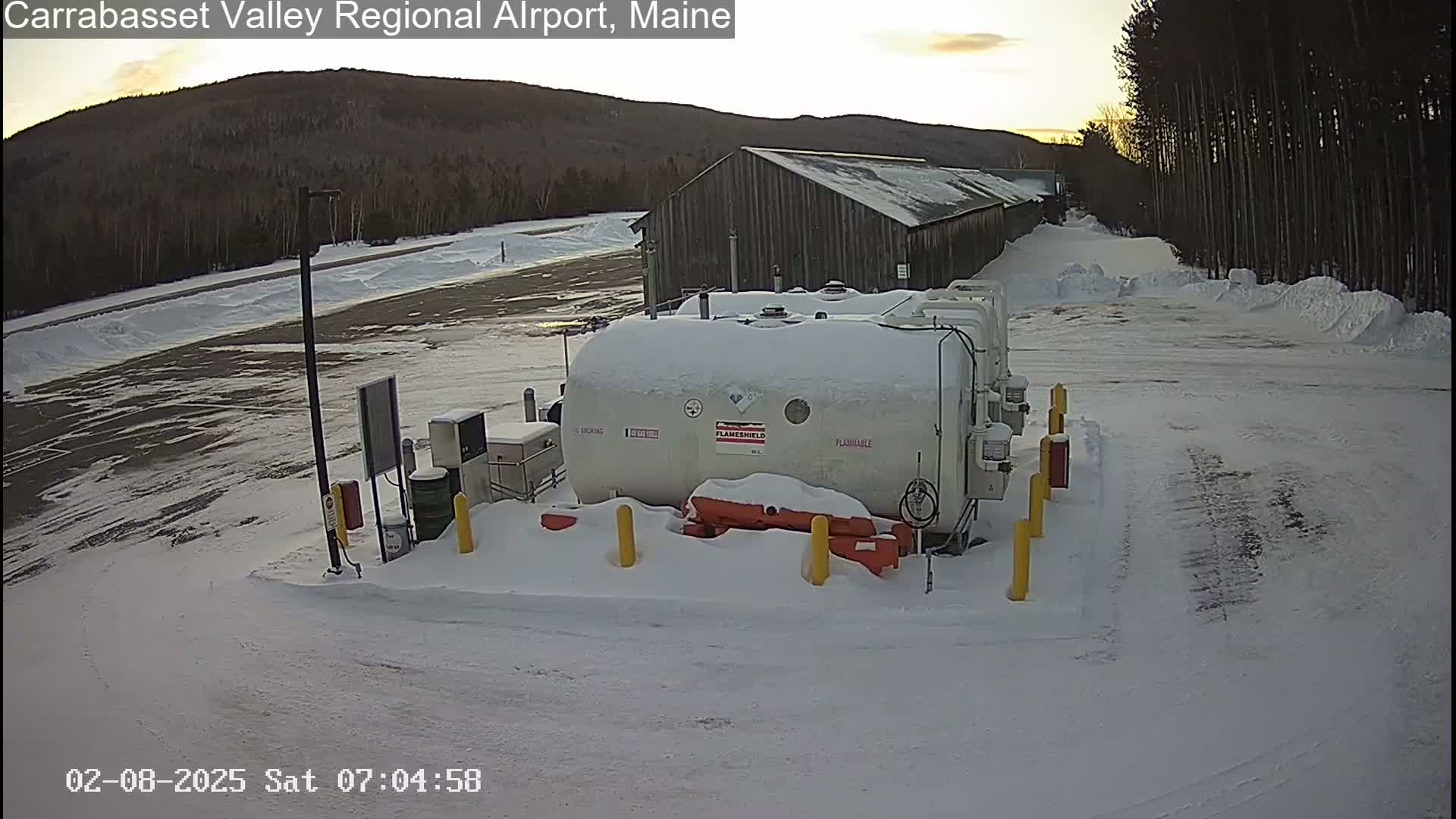 A snow-covered fuel storage area with a large, long building in the background during a sunrise.